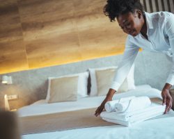 Eine Frau mit weisser Bluse legt Handtuecher auf ein gemachten Bett in einem Hotelzimmer