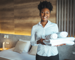 Frau mit einer weissen Bluse traegt Handtuecher auf dem Arm und steht in einem aufgeraeumten Hotelzimmer